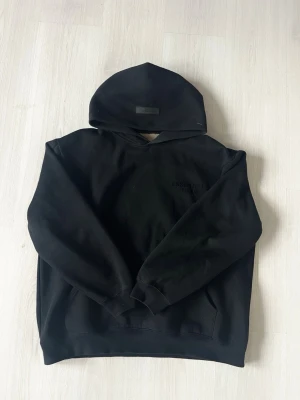 Svart Essentials Fear of God hoodie - Säljer en svart hoodie från Essentials Fear of God med diskret logga på bröstet och stor text på ryggen. Hoodien har klassisk passform, huva och ribbade muddar. Perfekt för dig som gillar streetwear och vill ha en stilren look.