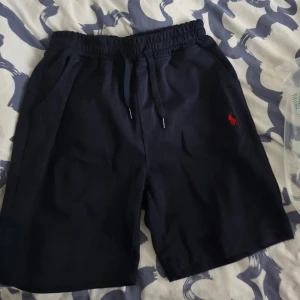 Mörkblå shorts med snörning - Snygga mörkblå shorts med elastisk midja och snörning framtill. Shortsen har två sidofickor och en liten röd logga broderad på vänster ben. Perfekta för sommardagar och avslappnade tillfällen.