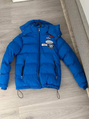 Blå pufferjacka med patches - Säljer en blå pufferjacka med huva och dragkedja framtill. Jackan har flera coola patches på bröstet och två fickor med dragkedja. Justerbar nederkant med snören och quiltad design för extra värme. Perfekt för kalla vinterdagar.