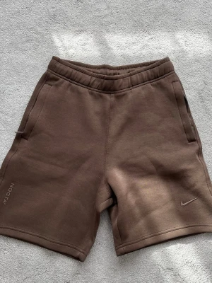 Bruna NOCTA x Nike shorts - Säljer ett par bruna shorts från NOCTA x Nike med elastisk midja och två sidofickor. Shortsen har NOCTA-logga på vänster ben och en liten Nike Swoosh på höger ben. Tillverkade i mjukt material som passar perfekt till sommaren.