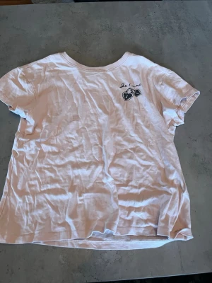 Rosa t-shirt från Kappahl med tryck - Rosa t-shirt från Kappahl med korta ärmar och ett litet tryck med texten 'Je t'aime' och två hjärtan på bröstet. Klassisk rund halsringning och mjukt bomullsmaterial som känns skönt mot huden. Perfekt till jeans eller shorts.