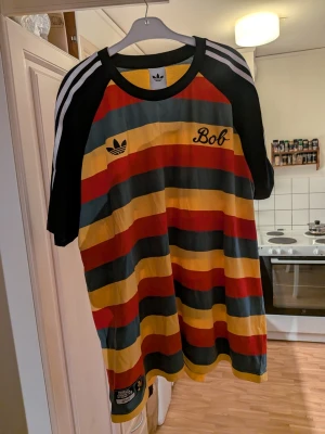 Randig Adidas Bob Marley t-shirt - Färgglad t-shirt från Adidas med breda ränder i rött, gult, grönt och svart. På bröstet står det 'Bob' och Adidas-loggan, och på nederdelen finns en patch med Bob Marley och texten 'Football is Freedom'. Klassisk Adidas-design med vita ränder på axlarna. Storlek XXXL.