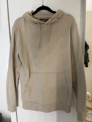 Beige hoodie med magficka och huva - Säljer en stilren beige hoodie med klassisk magficka och dragsko i huvan. Hoodien har långa ärmar och ribbade muddar vid ärmslut och nederkant. Perfekt för en avslappnad och enkel look.