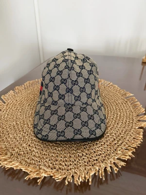 Beige keps med GG-mönster från Gucci - Snygg beige keps från Gucci med klassiskt GG-monogram i mörkblått över hela kepsen. På sidan finns en ikonisk röd och marinblå rand. Kepsen har böjd skärm och är tillverkad i canvasmaterial med svarta detaljer. Pris kan diskuteras.