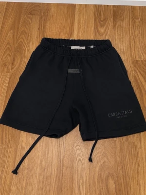 Svarta shorts från Essentials - Säljer ett par svarta shorts från Essentials med dragsko i midjan och diskret logga på benet. Sköna och stilrena, perfekta för chill dagar eller träning. Tillverkade i mjukt material med relaxed fit och elastisk midja.