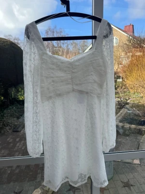 Helt ny vit spetsklänning - Supersöt vit kortklänning i spets med långärmade transparenta ärmar. Klänningen har diskret blommigt mönster i spetsen och dragkedja i ryggen. Det var en eventuell studentklänning som inte användes. Lapp kvar. 