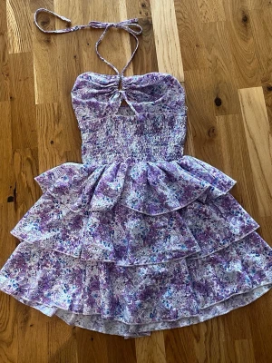 Lila blommig volangklänning - Supersöt kort klänning med lila och vita blommor, volangkjol i flera lager och smockad överdel. Klänningen har halterneck med knyt och en liten cut-out framtill. 