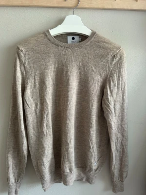 Beige tröja från NN07 - Snygg beige tröja från NN07 i storlek M. Tröjan har rund halsringning, långa ärmar och är tillverkad i ett tunt, mjukt material som passar perfekt för lager på lager. Enkel och stilren design som funkar till det mesta. Fått den av föräldrarna i present för ett tag sedan.