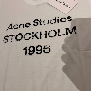 Vit Acne Studios t-shirt Stockholm - Säljer en vit t-shirt från Acne Studios med svart tryck på bröstet där det står 'Acne Studios STOCKHOLM 1996'. T-shirten har rund hals och korta ärmar. Perfekt för dig som gillar minimalistisk och stilren design.