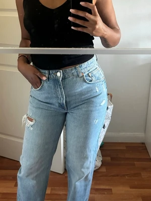 Ljusblå raka jeans med slitningar - Säljer ett par ljusblå högmidjade jeans med raka ben och trendiga slitningar på låret. Gjort i fint denimtyg. Perfekta för en avslappnad casual stil men går snabbt att klä upp till mer festlig outfit.