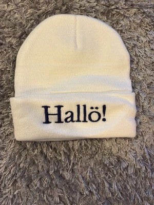 Hallå mössa - Säljer en vit ribbstickad mössa med uppvikt kant och svart broderad text 'Hallö!' framtill. Mössan är mjuk och stretchig, perfekt för att hålla värmen och ge en clean look. Passar dig som vill ha en enkel men personlig accessoar. Köpte men ångrade mig så använt typ en gång.
