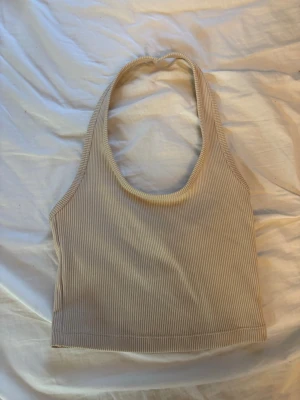 Beige ribbad linnetopp - Säljer en beige ribbad linnetopp med halterneck och croppad passform. Toppen är ärmlös och har en mjuk, stretchig känsla som passar perfekt till jeans eller kjol. Enkel och stilren design som är lätt att matcha.