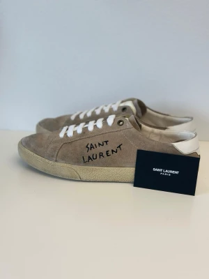 Saint Laurent beige mocka sneakers - Snygga beige sneakers från Saint Laurent i mocka med vit sula och vita skosnören. Handtextad logga på sidan och vit detalj vid hälen ger en clean och exklusiv look. Perfekta för dig som gillar stilrena och trendiga skor.