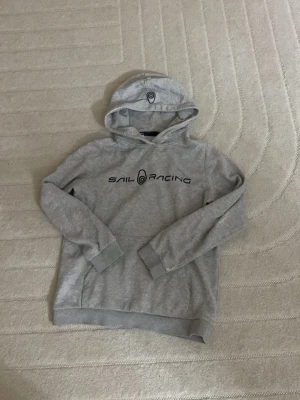 Grå hoodie från Sail Racing - Snygg grå hoodie från Sail Racing med svart logga och text på bröstet samt en broderad detalj på huvan. Tillverkad i mjukt bomullsmaterial och har klassisk känguruficka framtill. Perfekt för en avslappnad och sportig stil.