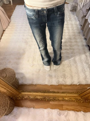 Blå bootcut jeans 25/30 - Säljer ett par blå bootcut jeans med snygga slitningar och ljusa tvättade partier framtill. Jeansen har låg midja, klassiska fem fickor och vita sömmar. Materialet är denim i bomull. Perfekta för en avslappnad och trendig look.