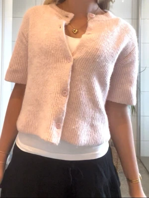 Ljusrosa kortärmad kofta  - Supersöt ljusrosa kofta med ribbad struktur och korta ärmar. Koftan har rund halsringning och rosa knappar. Den är i super fint skick!