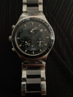 Stilren kronografklocka i silver - Snygg kronografklocka med svart urtavla och flera små urtavlor för extra funktioner. Klockan har en robust länkarmband i silverfärgad metall och markerad bezel. Passar perfekt för dig som gillar en klassisk men modern look.