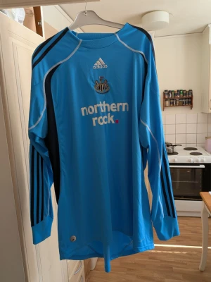 Blå Newcastle långärmad tröja Adidas - Säljer en blå långärmad Newcastle United tröja från Adidas med svarta och vita detaljer. Tröjan har klubbmärke och sponsortryck på bröstet samt klassiska Adidas-ränder på ärmarna. Tillverkad i funktionsmaterial, perfekt för fotbollsträning. Storlek  XXL