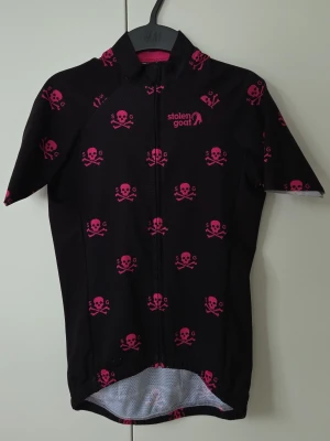 Svart cykeltröja med rosa dödskallar - Snygg svart cykeltröja med rosa dödskallar från Stolen Goat, hel dragkedja framtill och korta ärmar. Tröjan har 3 praktiska fickor på ryggen + en vattentät sidoficka med dragkedja. Tillverkad i ett lätt och ventilerande material som passar perfekt för cykling.