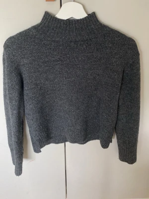 Mörkgrå stickad tröja med hög krage - En enkel och stilren mörkgrå stickad tröja med hög ribbad krage och långa ärmar. Tröjan har en rak passform och ribbade muddar vid ärmslut och nederkant. Perfekt för kalla dagar och lätt att matcha med olika outfits.