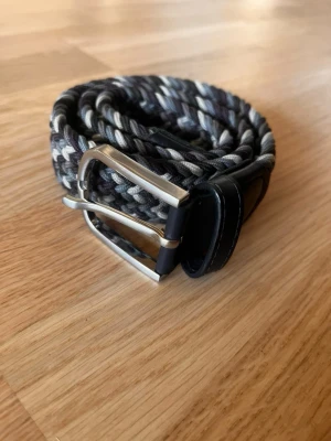 Onewear - flätat bälte - Snyggt flätat bälte i svart, grått och vitt med silverfärgat spänne. Bältet har en cool, vävd look och passar perfekt till jeans eller chinos. Detaljer i svart konstläder vid spännet ger en stilren touch.