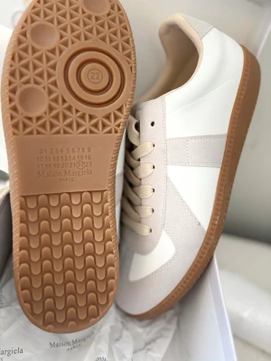 Maison Margiela vita och beige sneakers - Maison Margiela sneakers i vitt och beige med klassisk låg siluett. Skorna har ovandel i slätt vitt läder och beige mockadetaljer, samt beige snörning och brun gummisula. Perfekta för en clean och stilren look.