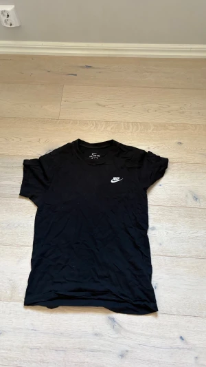 Svart Nike t-shirt med logga - En klassisk svart t-shirt från Nike med vit broderad logga på bröstet. T-shirten har rund halsringning och korta ärmar. Tillverkad i mjuk bomull för en skön och avslappnad stil.