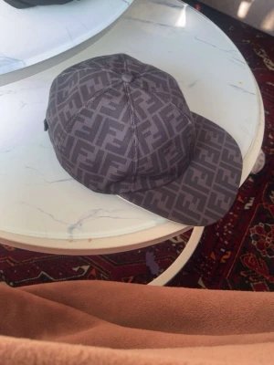 Mörkgrå Fendi keps med FF-mönster - Säljer en mörkgrå keps från Fendi med det klassiska FF-logomönstret över hela kepsen. Kepsen har rak skärm och justerbar baksida. Materialet är slitstarkt tyg som känns lyxigt och trendigt. Perfekt för dig som vill ha en statement-accessoar.