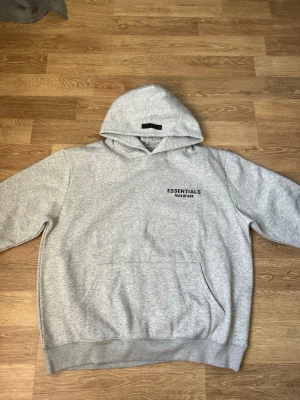 Grå Essentials Fear of God hoodie - Snygg grå hoodie från Essentials Fear of God med svart logga på bröstet. Klassisk passform med stor huva och magficka. Tillverkad i mjuk bomullsblandning som känns riktigt skön mot huden. Perfekt för en avslappnad streetwear-look.