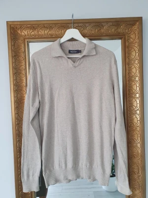 Beige stickad tröja med krage - Stilren beige stickad tröja med klassisk krage och v-ringning. Tröjan har långa ärmar och ribbade muddar vid ärmslut och nederkant. Perfekt för en avslappnad men ändå snygg look.