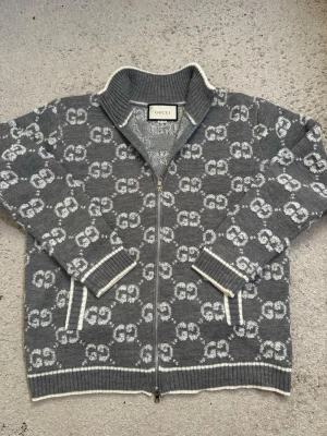 Grå ziptröja med GG-mönster från Gucci - Snygg grå ziptröja från Gucci med vitt GG-mönster över hela plagget. Tröjan har ribbade muddar, hög krage och vita detaljer längs kanterna. Perfekt för dig som vill ha en exklusiv och trendig look.