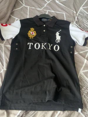 Svart piké Tokyo Polo Ralph Lauren - Snygg svart pikétröja från Polo Ralph Lauren med vita ärmar, broderad logga och Tokyo-tryck på bröstet. Slim fit-modell med krage och knappar framtill. Detaljerad brodyr i guld och vitt på vänster sida. Perfekt för en stilren och sportig look.