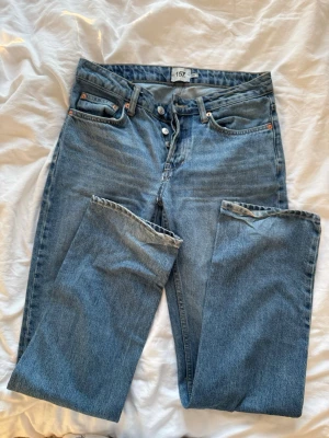 Blå raka jeans med hög midja - Klassiska blå jeans med rak passform och medel hög midja. Jeansen har knappgylf, fem fickor och subtila slitningar vid benslut. Perfekta för en avslappnad och trendig look.