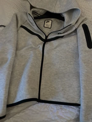 Ljusgrå zip hoodie från Nike - Snygg ljusgrå hoodie från Nike med hel dragkedja och svarta detaljer längs dragkedjan och ärmarna. Hoodien har en klassisk huva och sportig look, perfekt för chill dagar eller träning. Tillverkad i mjukt material för extra komfort.