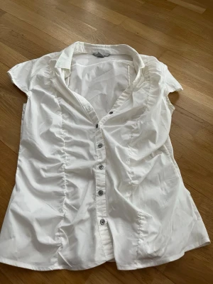 Vit kortärmad blus från H&M - Klassisk vit blus från H&M med korta ärmar och knappar framtill. Blusen har lätt rynkade sidor och en snygg krage. Tillverkad i ett tunt och luftigt material som passar perfekt till många olika stilar.