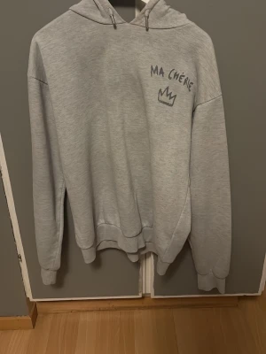 Oversize Grå hoodie från Gina Tricot - Ljusgrå hoodie från Gina Tricot med trycket 'MA CHÉRIE' och en krona på bröstet. Hoodien har huva med dragsko och ribbade muddar vid ärmar och nederkant. Mjuk insida för extra komfort, perfekt för chill dagar.