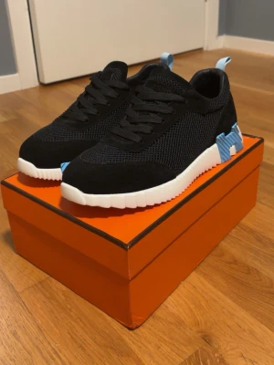 Svarta mesh sneakers med blå detalj - Snygga svarta sneakers med ovandel i mesh och mocka, vit räfflad sula och blå detaljer vid hälen. Skorna har svarta skosnören och en modern, sportig look. Perfekta för dig som gillar stilrena men ändå unika sneakers.