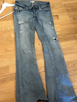 Blå slitna bootcut jeans - Säljer ett par ljusblå bootcut jeans med slitningar och hål på benen. Jeansen har klassisk femficksmodell, låg midja och råa detaljer för en trendig look. Perfekta för dig som gillar en avslappnad och edgy stil.