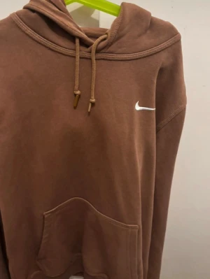 Brun hoodie med vit Nike-logga - Snygg brun hoodie från Nike med vit broderad Swoosh på bröstet. Hoodien har justerbar dragsko i huvan och en stor magficka framtill. Tillverkad i mjukt material som känns skönt mot huden. Perfekt för en avslappnad och trendig look.