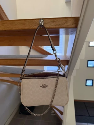Vit axelremsväska från Coach - Snygg vit handväska från Coach med klassiskt monogrammönster. Väskan har både en kort axelrem i läder och en dekorativ silverfärgad kedja. Dragkedja upptill och ljusbrunt innerfoder. Perfekt storlek för det viktigaste!