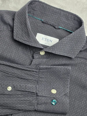 Eton Chelsea skjorta mörkgrå storlek M - Mörkgrå skjorta från Eton i Slim-passform och modellen Chelsea. Diskret vävt ton-i-ton-mönster som ger skjortan ett stilrent och elegant uttryck. Tillverkad i 100% bomull med ett tunt och luftigt tyg som gör den särskilt lämpad för vår- och sommarsäsongen.  Storlek M / 40 Slim Fit  Mycket fint skick.  Mått Bröstbredd: ca 52 cm Plagglängd: ca 70 cm Axelbredd: ca 42 cm Ärmlängd: ca 65 cm  Material 100% bomull  Cut away-krage, bröstficka och diskret ton-i-ton-mönster.