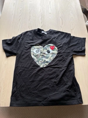 Svart t-shirt med camohjärta comme des garcons - Svart t-shirt från comme des garcons med ett stort hjärta i camouflage-mönster och ögon på bröstet. Ovanpå hjärtat sitter en liten röd hjärtdetalj med ögon. Klassisk passform och rund hals. Perfekt för dig som vill sticka ut med en unik design.