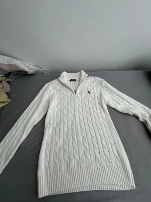 Stickad polo tröja - Kabelstickad vit tröja från Ralph Lauren med lång ärm och ribbade muddar. Tröjan har en liten krage med dragkedja och den klassiska broderade loggan på bröstet. Perfekt för dig som gillar en clean och tidlös stil. Nytt utanprislapp den är med kommer med helt nytt inte använd Den är M funkar S