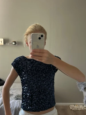 Mörkblå paljettopp med kort ärm - Snygg mörkblå topp täckt med glittriga paljetter. Modellen har korta ärmar och en rak, croppad passform. Tyget är mjukt på insidan för skön känsla mot huden.