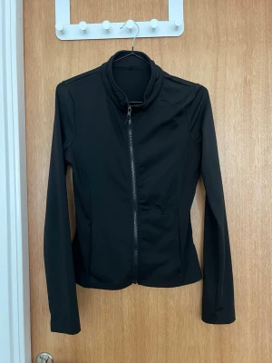 Svart sportjacka med dragkedja - Snygg svart sportjacka med hel dragkedja framtill och hög krage. Jackan har lång ärm och är tillverkad i ett stretchigt, lätt material som passar perfekt till träning eller löpning. Figurnära passform för en sportig look.