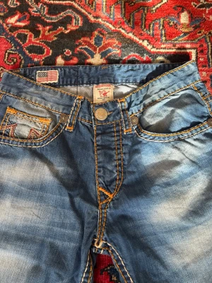 Blå True Religion jeans  - Säljer ett par blå jeans med tydliga kontrastsömmar i orange och slitna detaljer. Klassisk femficksmodell med knappgylf och coola detaljer på fickorna. Jeansen har en rak passform och är tillverkade i denim.