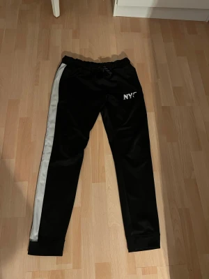 Byxor - A pair of black athletic pants with a white stripe running down the left leg and 'NYC' printed on the upper left thigh. The pants are laid flat on a light wooden floor.