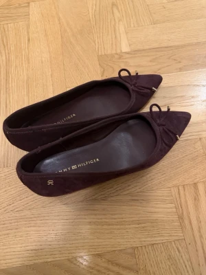 Vinröda ballerinaskor Tommy Hilfiger - Snygga vinröda ballerinaskor från Tommy Hilfiger i mockaimitation. Skorna har spetsig tå, dekorativ rosett framtill och små gulddetaljer. Perfekta för dig som vill ha en stilren och trendig look. I storlek 6M vilket motsvarar ca 36. Använda en gång