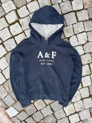 Mörkblå hoodie med teddyfoder A&F - Mysig mörkblå hoodie från A&F med vit text 'A&F New York Est. 1892' på bröstet. Hoodien har en stor huva med mjukt teddyfoder och träknappar vid halsen. Praktisk magficka framtill och ribbade muddar vid ärmslut och nederkant. Storlek L men sitter som S