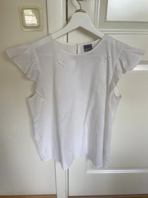 Vit blus från Ginatricot i storlek 38 - Söt vit blus med broderade blommor och korta volangärmar. Toppen har en luftig passform och är tillverkad i bomull, vilket gör den perfekt för varma dagar. Blusen har fina detaljer med spetsliknande broderier som ger en romantisk känsla.